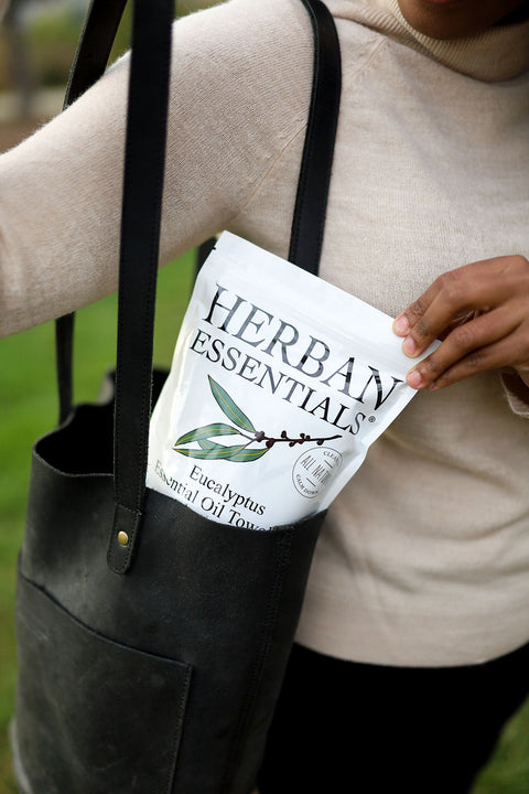 Woman placing a pouch of Herban Essentials Eucalyptus Essential Oil Towelettes into a black leather tote bag, highlighting their portability and refreshing eucalyptus benefits for travel, work, and everyday use.