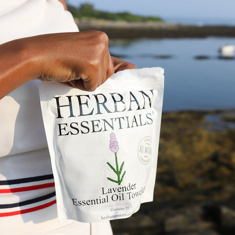 Close-up of a woman holding a pouch of Herban Essentials Lavender Essential Oil Towelettes by the water, highlighting their portability and refreshing aromatherapy benefits for travel, outdoor days, and self-care.