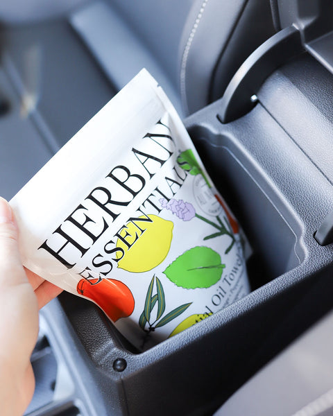 Hand placing a pouch of Herban Essentials Essential Oil Towelettes into a car console, showing their portability for everyday hygiene and aromatherapy.