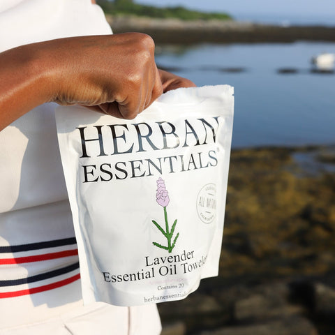 Person holding a Herban Essentials lavender essential oil product by a waterfront.