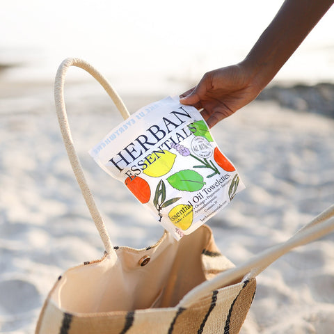 Person holding a 'Herban Essentials' product over a striped bag on a beach.