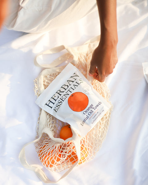 Herban Essentials Orange Essential Oil Towelettes in a mesh tote with oranges, on a white blanket.