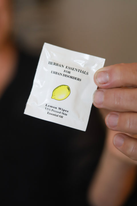 Hand holding a packet of original 'Herban Essentials' lemon wipes against a blurred background
